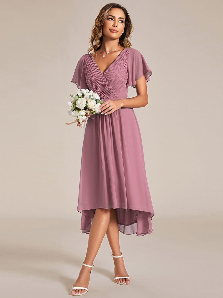 Model in the mauve-pink chiffon dress holding a small bouquet of white flowers, glancing to the side in white heels. 60