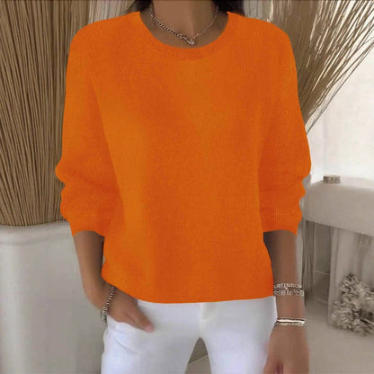 Model wears the round-neck knit jumper in bright orange against a neutral interior with dried grasses 11