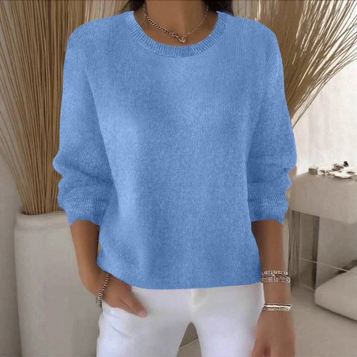 Model wears the round-neck knit jumper in light blue, standing by a vase of dried grasses 13