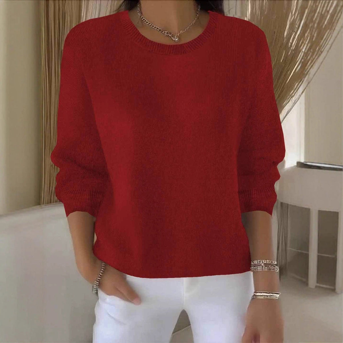 Model wears the round-neck knit jumper in deep red with white trousers and silver jewellery 1