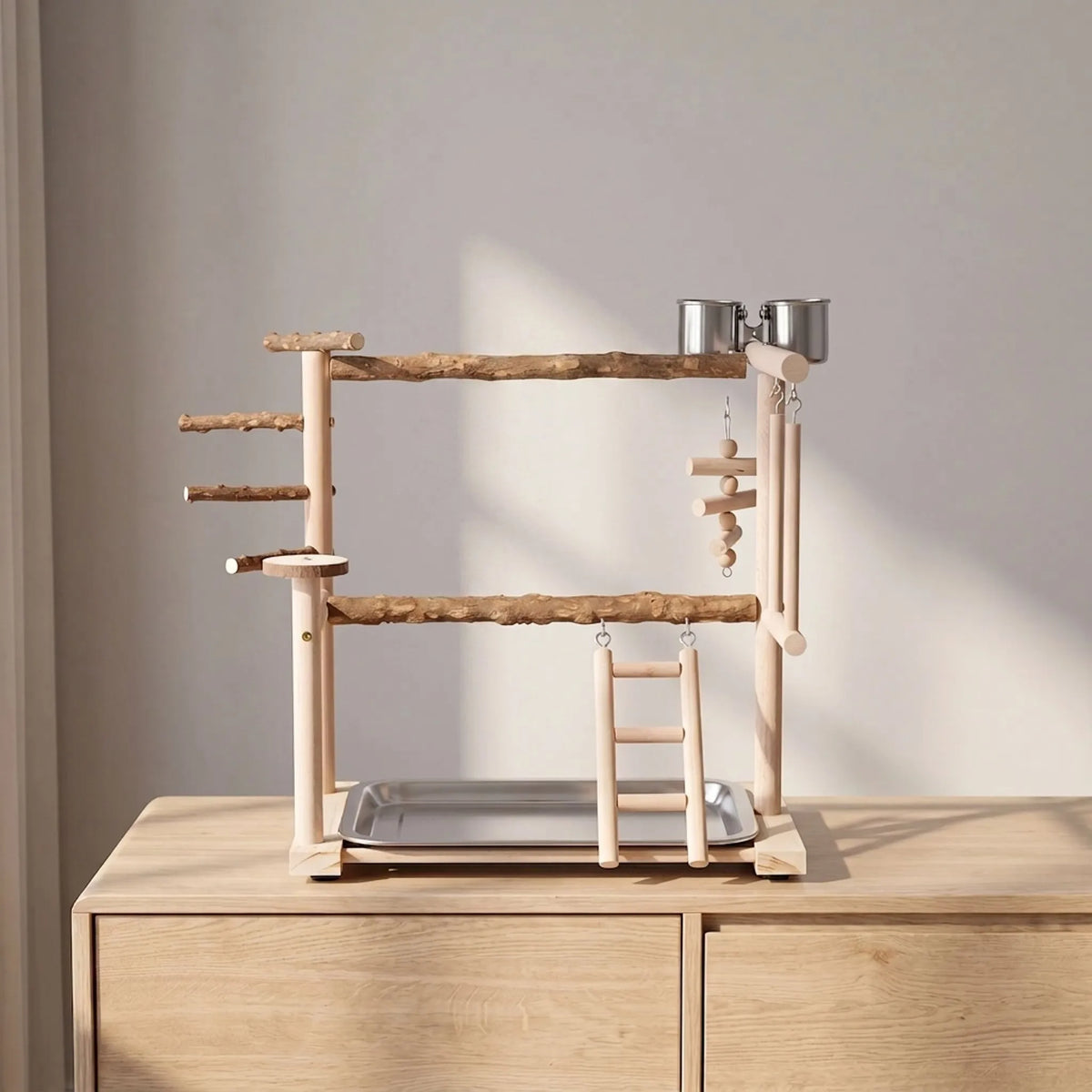 Wooden bird playground with two branch perches, ladder, swings and steel cups, standing on a pale wooden sideboard against a wall. 3