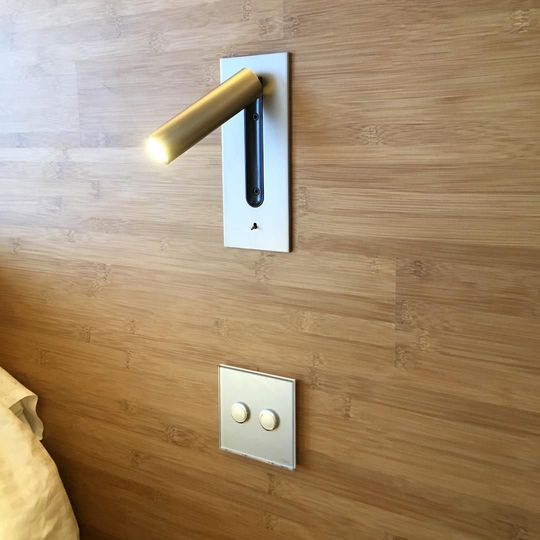 Brass-toned LED tube extended horizontally from a silver plate on a bamboo wall, above a two-button switch 29