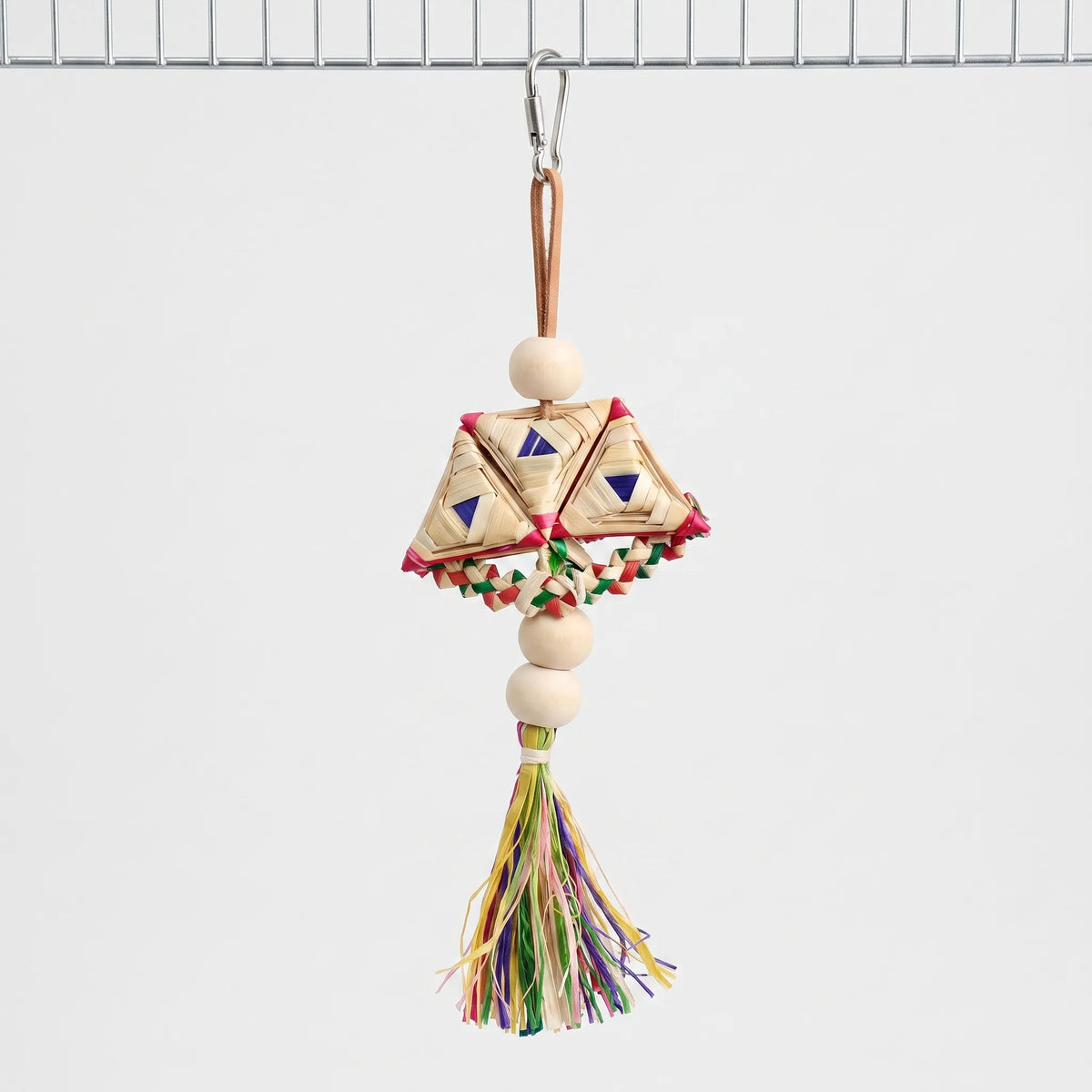 Woven straw toy with wooden beads and a colourful raffia tassel hanging from a steel clip on a wire cage panel. 1