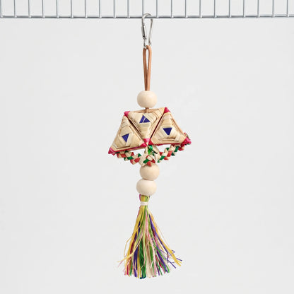 Woven straw toy with wooden beads and a colourful raffia tassel hanging from a steel clip on a wire cage panel. 1