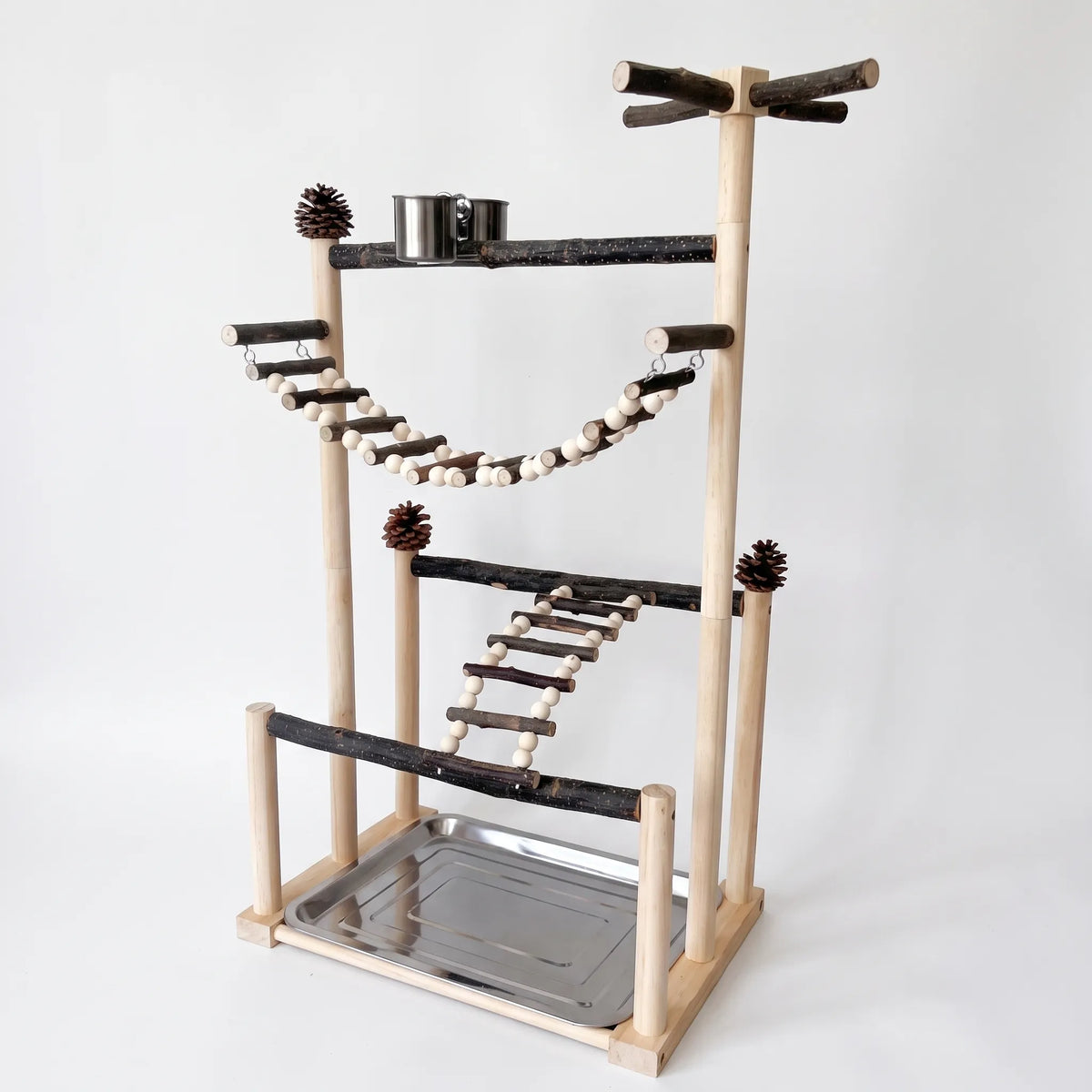 Wooden bird play stand with bark perches, a beaded rope bridge, a ladder, pine cones, a steel food cup and a base tray, on plain white. 1