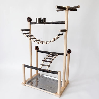 Wooden bird play stand with bark perches, a beaded rope bridge, a ladder, pine cones, a steel food cup and a base tray, on plain white. 1