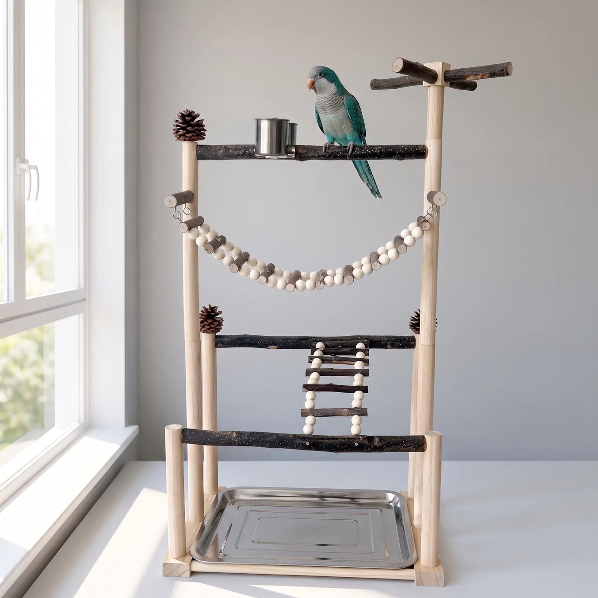 The same bird play stand beside a bright window, with a blue-green parrot perched on the top bark branch next to the steel cup. 2