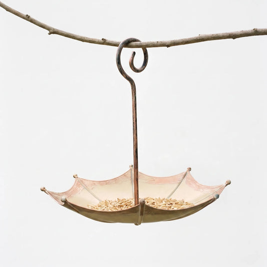 Upturned-umbrella metal feeder holding seed, its scrolled copper-toned handle hooked over a bare branch against a plain pale background. 1