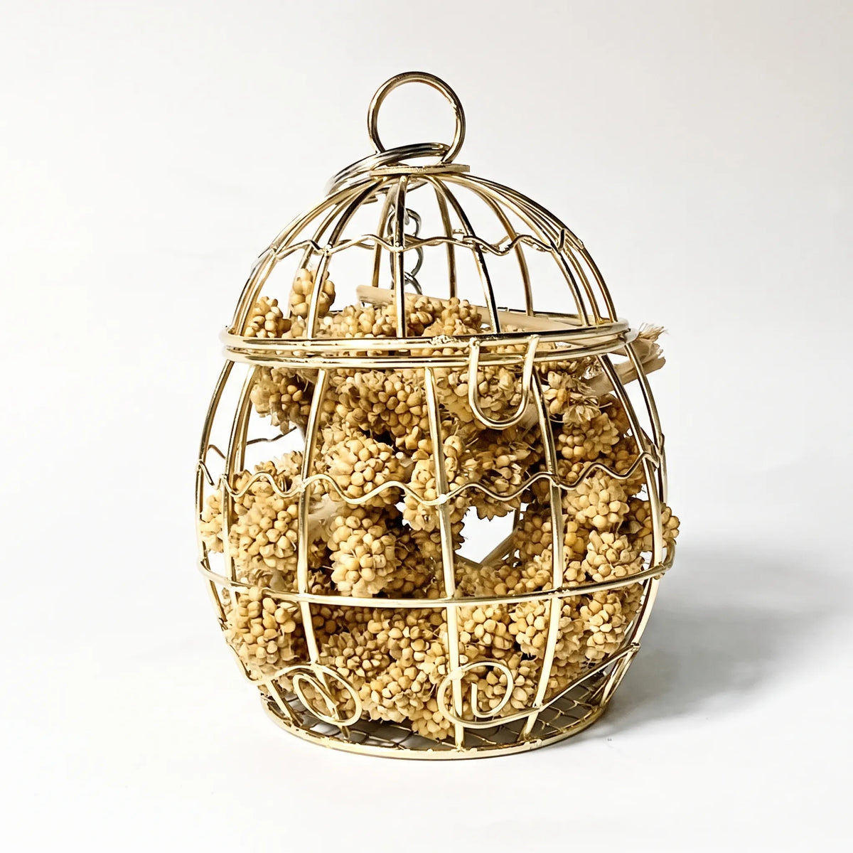 Egg-shaped gold wire cage with a ring on top, packed with dried millet sprays, shown against a plain white background. 1
