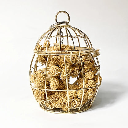 Egg-shaped gold wire cage with a ring on top, packed with dried millet sprays, shown against a plain white background. 1