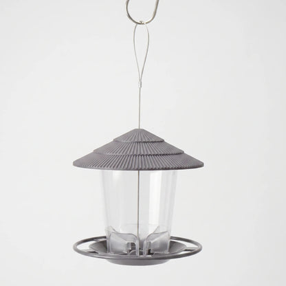 Grey hanging feeder with a ridged tiered roof, clear seed tube and round perch tray, hung from a wire loop on a hook. 1