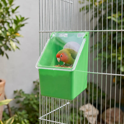 Feeder hanging on the wire of a cage outdoors, with a rosy-faced lovebird perched inside eating seed. 2