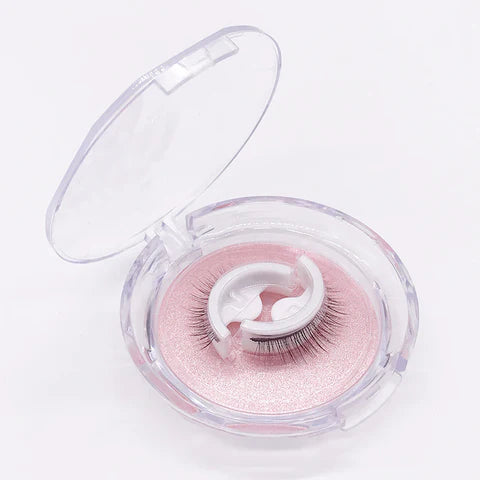 Open clear round case with a pair of self-adhesive lashes resting on white curved holders over a pink glittery tray. 1