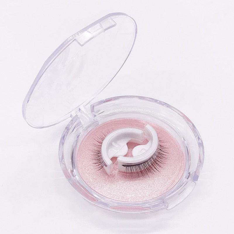 Open clear round case showing a pair of self-adhesive lashes on white curved holders over a pink glittery tray, on a white background. 6