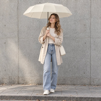 Woman in a beige coat and jeans stands against a concrete wall, holding the cream umbrella open overhead. 1