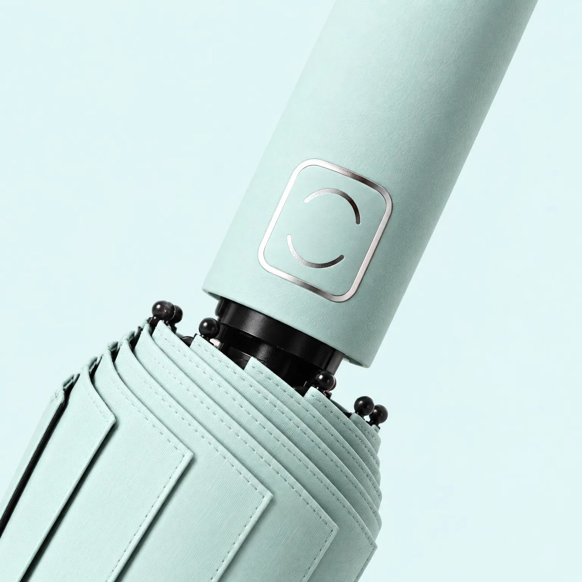 Close-up of a pale mint handle with a metallic square emblem and the folded canopy's stitched pleats and black rib tips. 5
