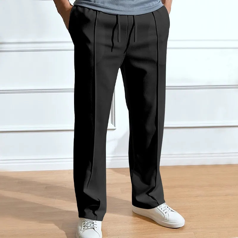 Model wearing the trousers in black, hands in pockets, drawstring waist and vertical front creases visible, paired with white trainers. 3