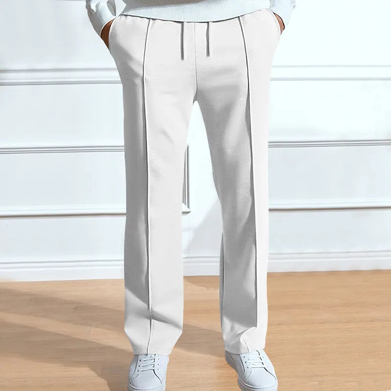 Front view of the trousers in white on a model, with drawstring waist, front pleat lines and hands in pockets, worn with white trainers. 4