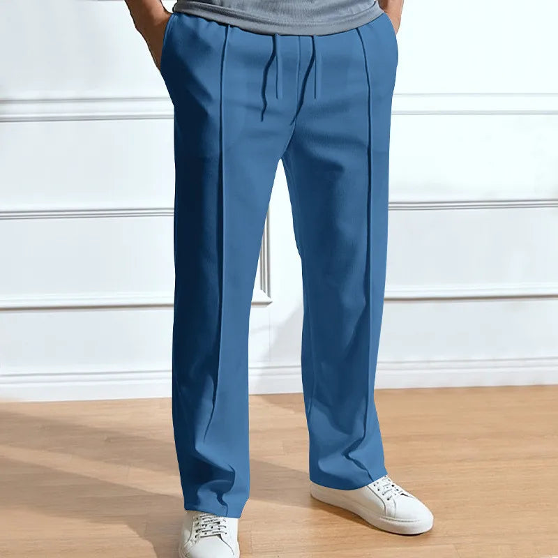 Model wearing the trousers in mid-blue, showing the drawstring waist and front creases, hands in pockets and white trainers. 5