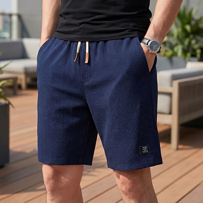 Model wearing navy blue waffle-textured shorts with a cream drawstring and brown tips, one hand in a pocket, on a terrace. 7
