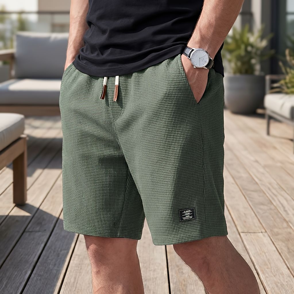 Model wearing green waffle-textured shorts with a cream drawstring and brown tips, one hand in a pocket, on a wooden deck. 5
