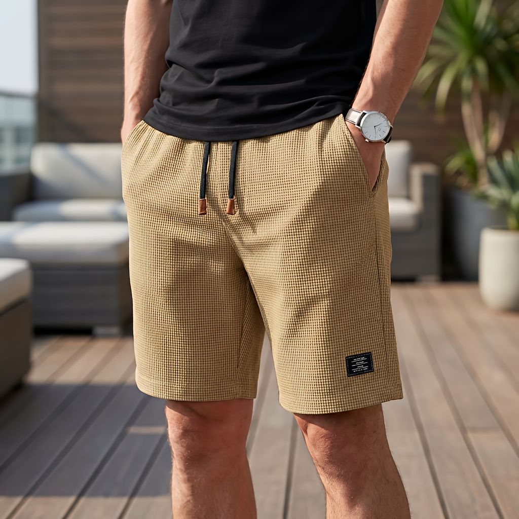 Model wearing sand-coloured waffle-textured shorts with black drawstring and brown tips, one hand in a pocket, on a sunny terrace. 4