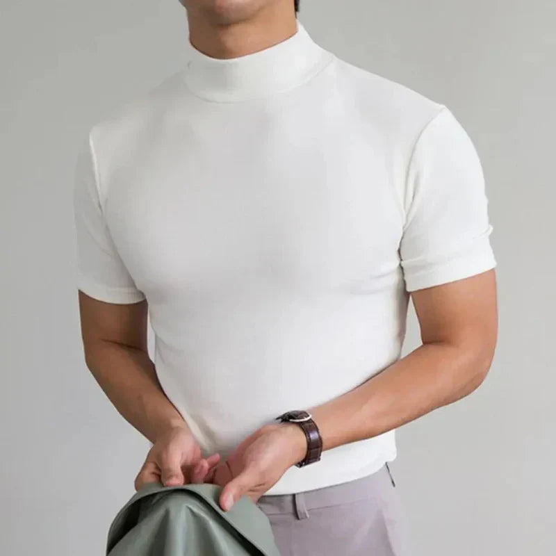 Model in the white high-neck t-shirt with a brown leather-strap watch, holding a green jacket in front of light trousers. 4