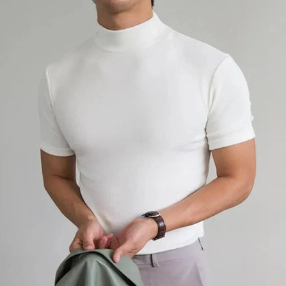 Model in the white high-neck t-shirt with a brown leather-strap watch, holding a green jacket in front of light trousers. 4