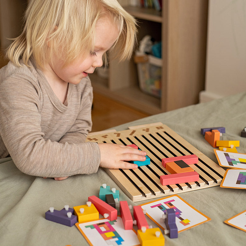 Fair-haired toddler in a grey top concentrating on fitting blocks into the slotted board, with loose pieces and pattern cards around. 4