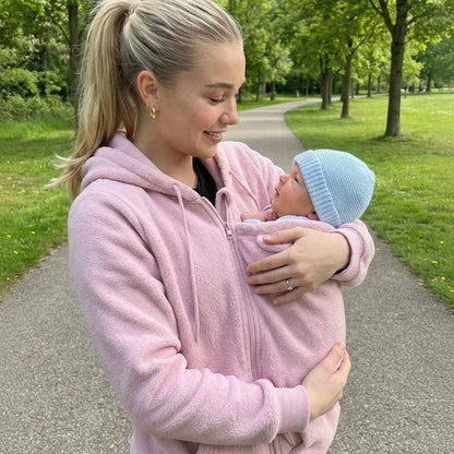 Woman in a soft pink fleece hoodie on a park path, gazing at her baby in a blue knitted hat. 4