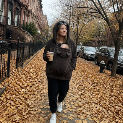 Woman in a dark brown sherpa hoodie with the hood up, walking along a leafy street with a baby in the panel. 5