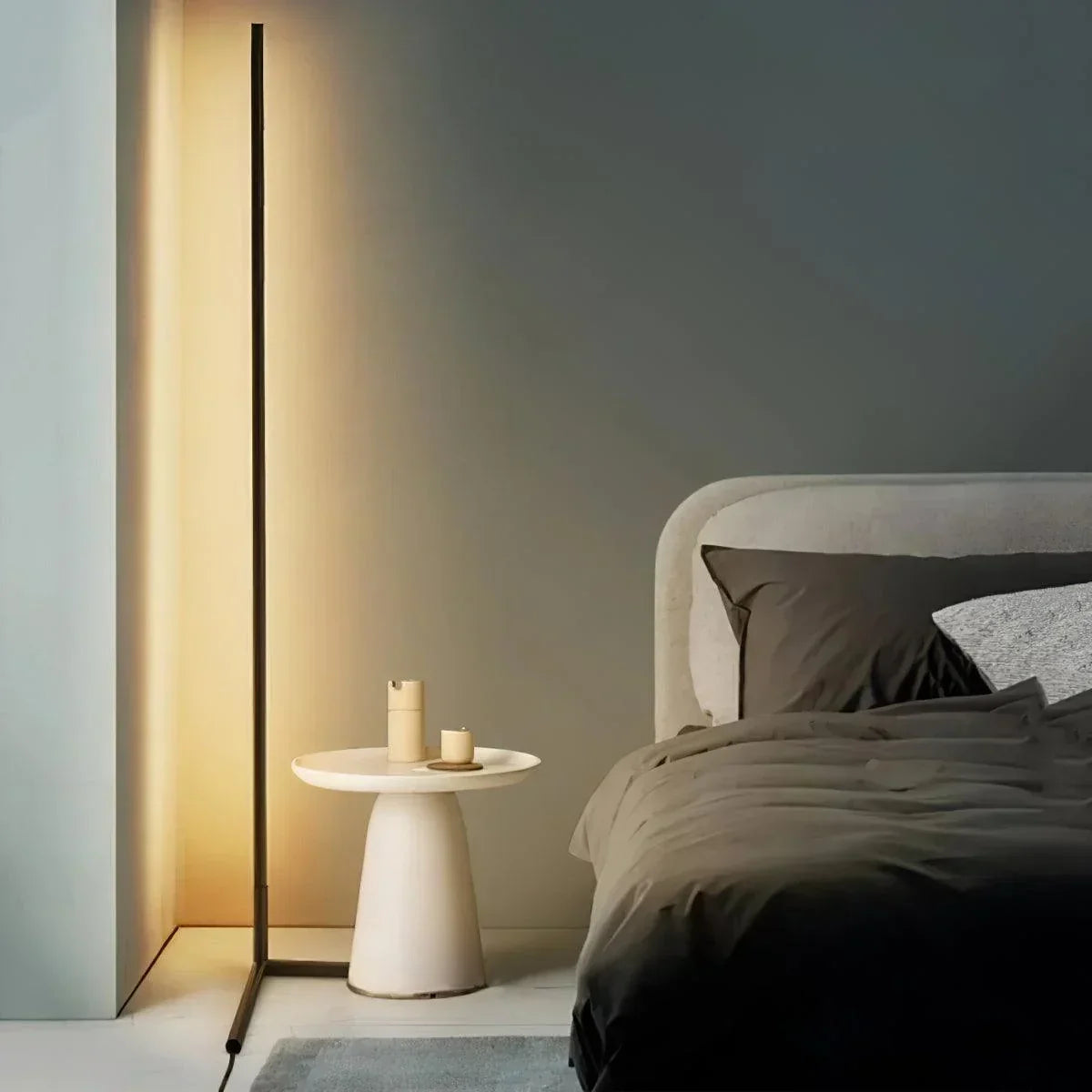 Slim black lamp glowing warm white beside a cream pedestal side table and a bed 1