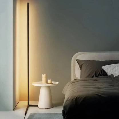 Slim black lamp glowing warm white beside a cream pedestal side table and a bed 1