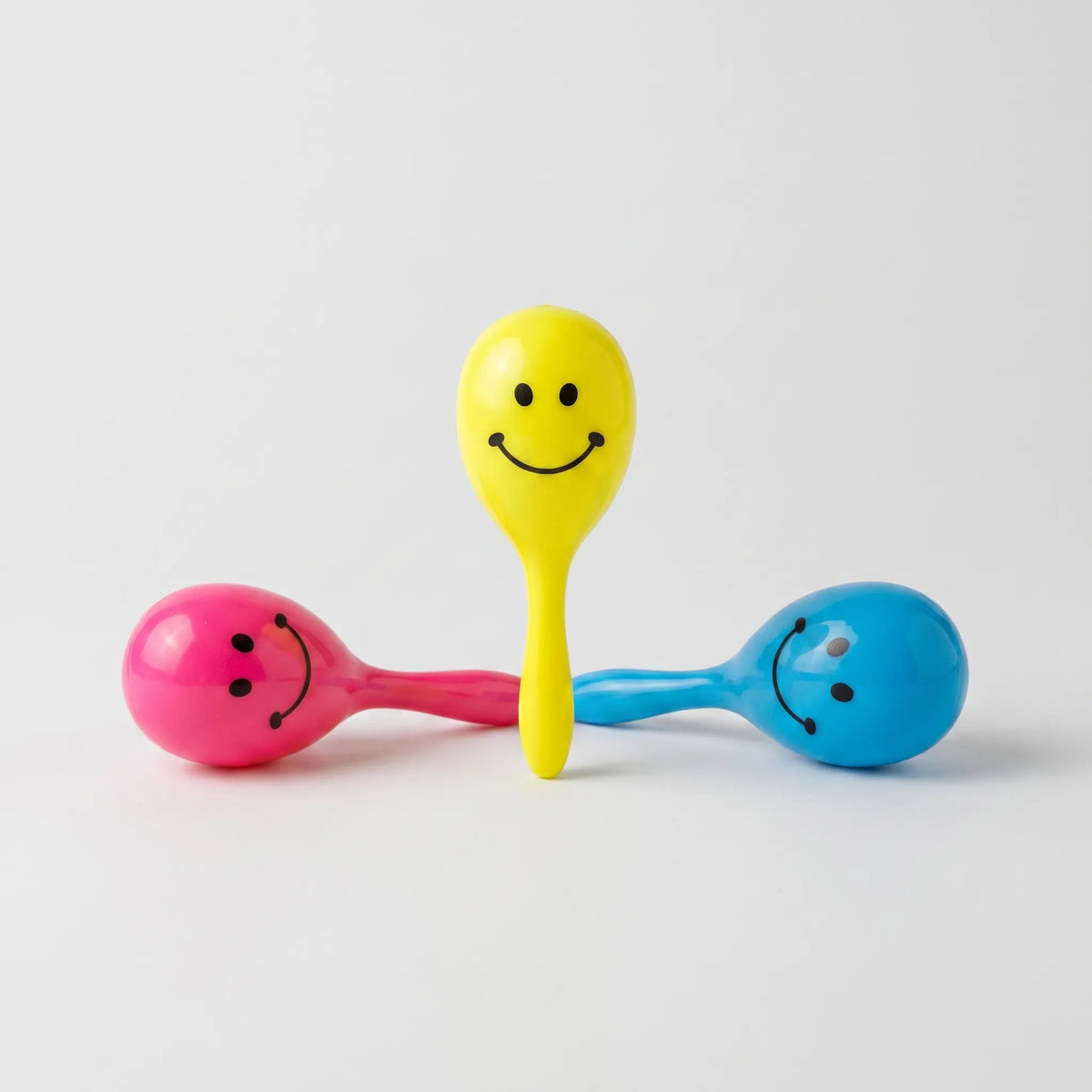 Three glossy plastic maracas with printed smiley faces in yellow, pink and blue, one standing upright between two lying down. 1