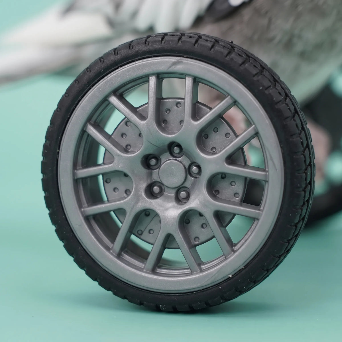 Close-up of one wheel showing its grey multi-spoke rim and black treaded rubber tyre against a turquoise background. 3