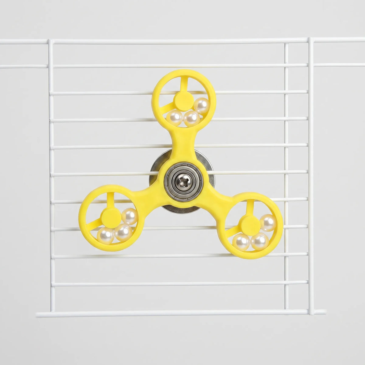 Front view of the yellow three-armed spinner toy fixed to white cage bars, with pearl-like beads in each ring. 1