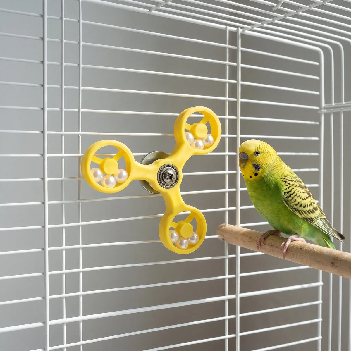 Yellow spinner toy attached to white cage bars while a green budgie sits on a wooden perch next to it. 3