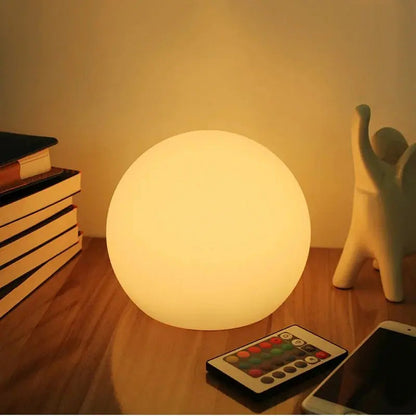 Glowing warm-white orb lamp on a wooden desk beside stacked books, a small figurine, a colour remote control and a smartphone. 1