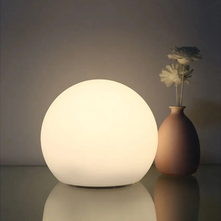 Softly lit white orb lamp on a reflective surface next to a rounded vase holding two pale flowers. 2