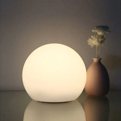 Softly lit white orb lamp on a reflective surface next to a rounded vase holding two pale flowers. 2