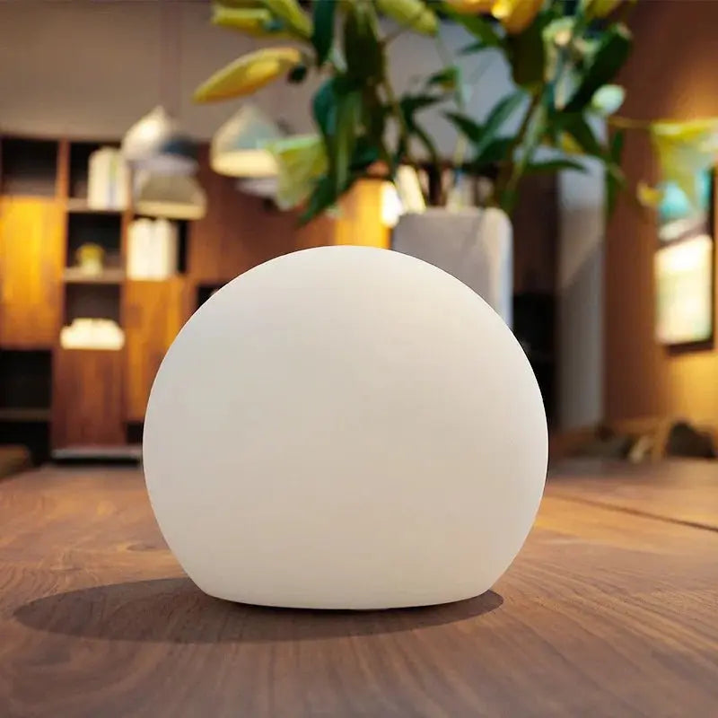 Unlit white spherical lamp standing on a wooden table, with a leafy plant and shelving blurred in the background. 3