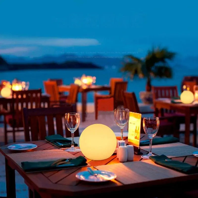 Orb lamp glowing yellow on an outdoor restaurant table set with glasses and plates, with a seaside view at dusk. 5