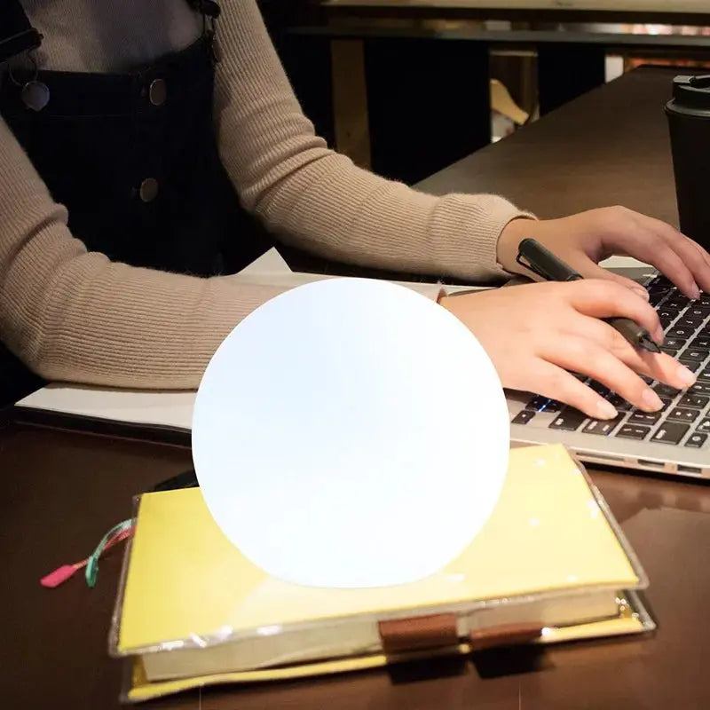 Bright white orb lamp resting on a closed notebook while a person in a beige jumper types on a laptop. 7