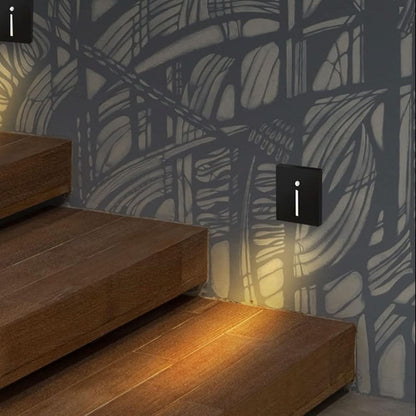 Two black slot-style wall lights glowing warmly beside wooden stairs against a patterned grey wall 2
