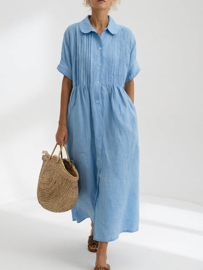 Model in the pale blue dress facing front, showing the full button placket, pintucked bodice and rounded collar, holding a straw tote. 2