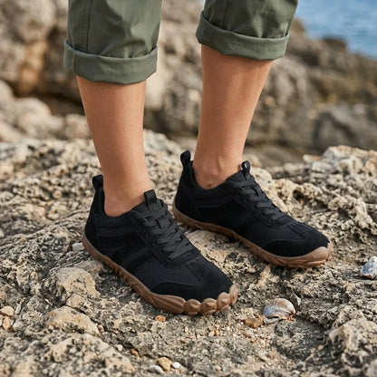 Black barefoot shoes worn on rocky shoreline with rolled olive trousers, tan soles and a shell nearby. 20