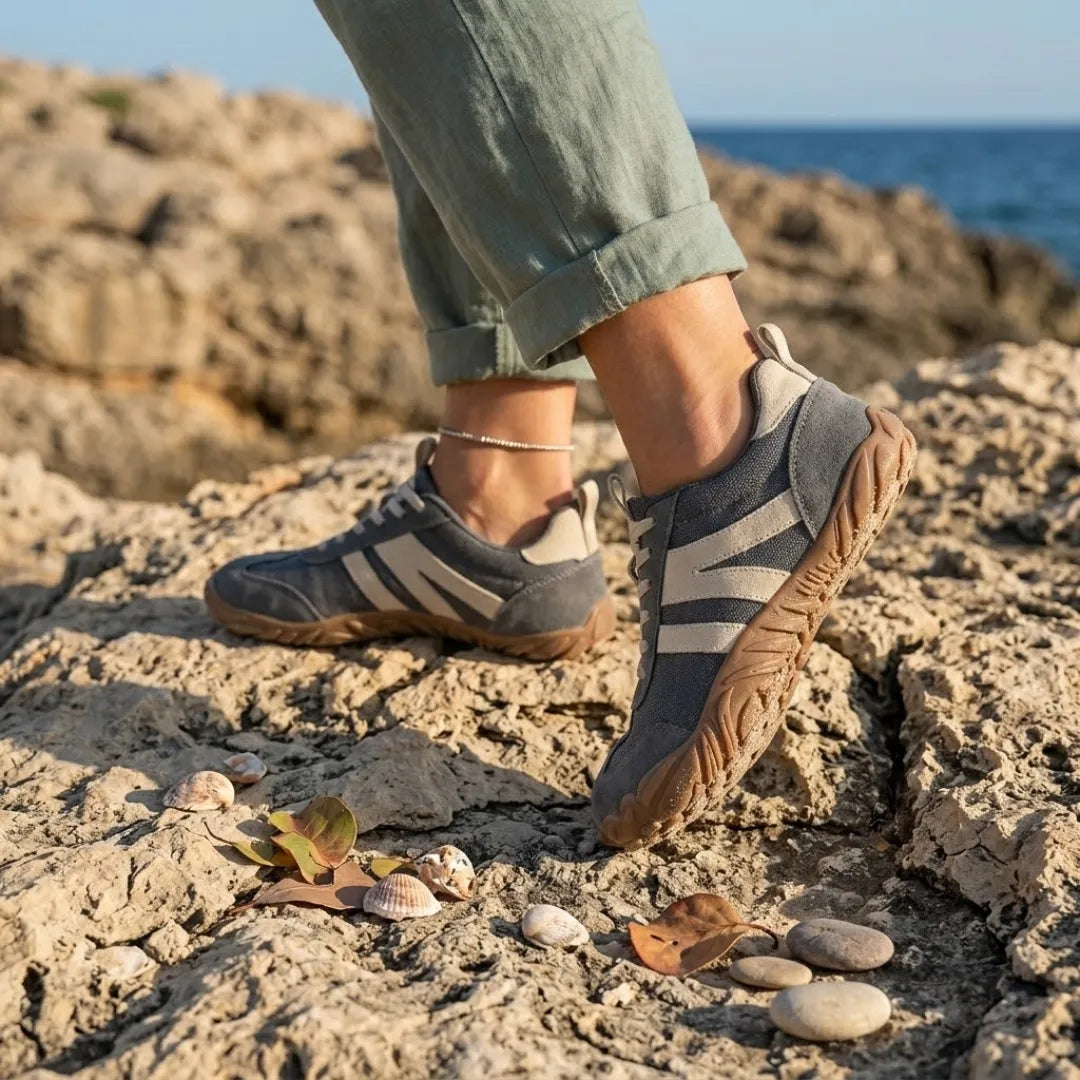 Grey barefoot shoes worn on rocky coastline with rolled green linen trousers, shells and pebbles beside them. 2