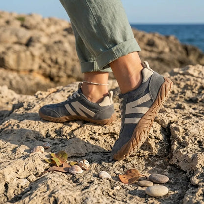 Grey barefoot shoes worn on rocky coastline with rolled green linen trousers, shells and pebbles beside them. 2