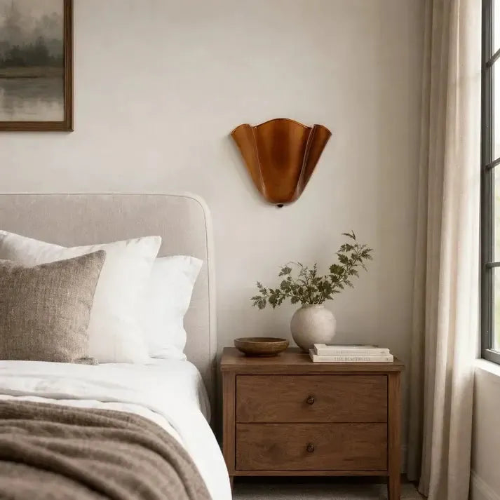Unlit amber-brown wavy sconce mounted above a wooden bedside chest with a vase, books and neutral linen bedding. 3