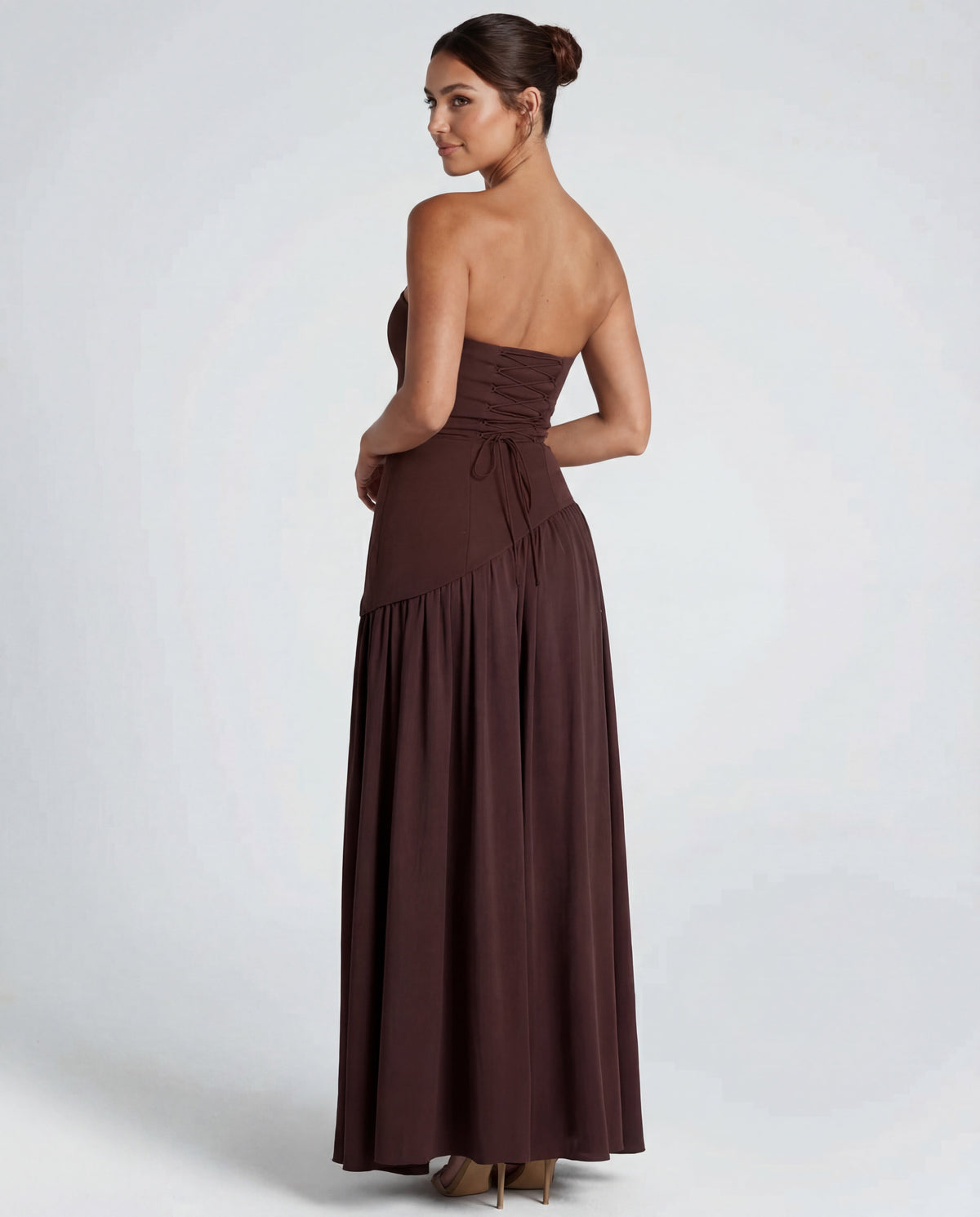 Back view of brown strapless maxi showing corset lacing at the waist and gathered skirt, model glancing over shoulder 2
